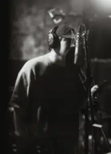 Wearing headphones and a cap, a person stands in front of a microphone in a recording studio, holding a mobile device and using SongTools to enhance their music production.